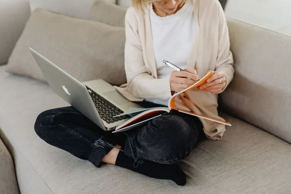 Une personne assise sur un canapé prend des notes à côté de son ordinateur portable.